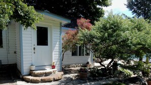 A small, single-story mobile home with a porch and steps leading to the door. There are potted plants and a small garden area with a tree and shrubs. - Beautiful Beach front cottage near Nelson, BC (Nelson)