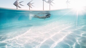 Une personne nageant sous l’eau dans une piscine aux eaux cristallines, dont la surface est illuminée par le soleil, avec une île lointaine où se dressent des éoliennes.