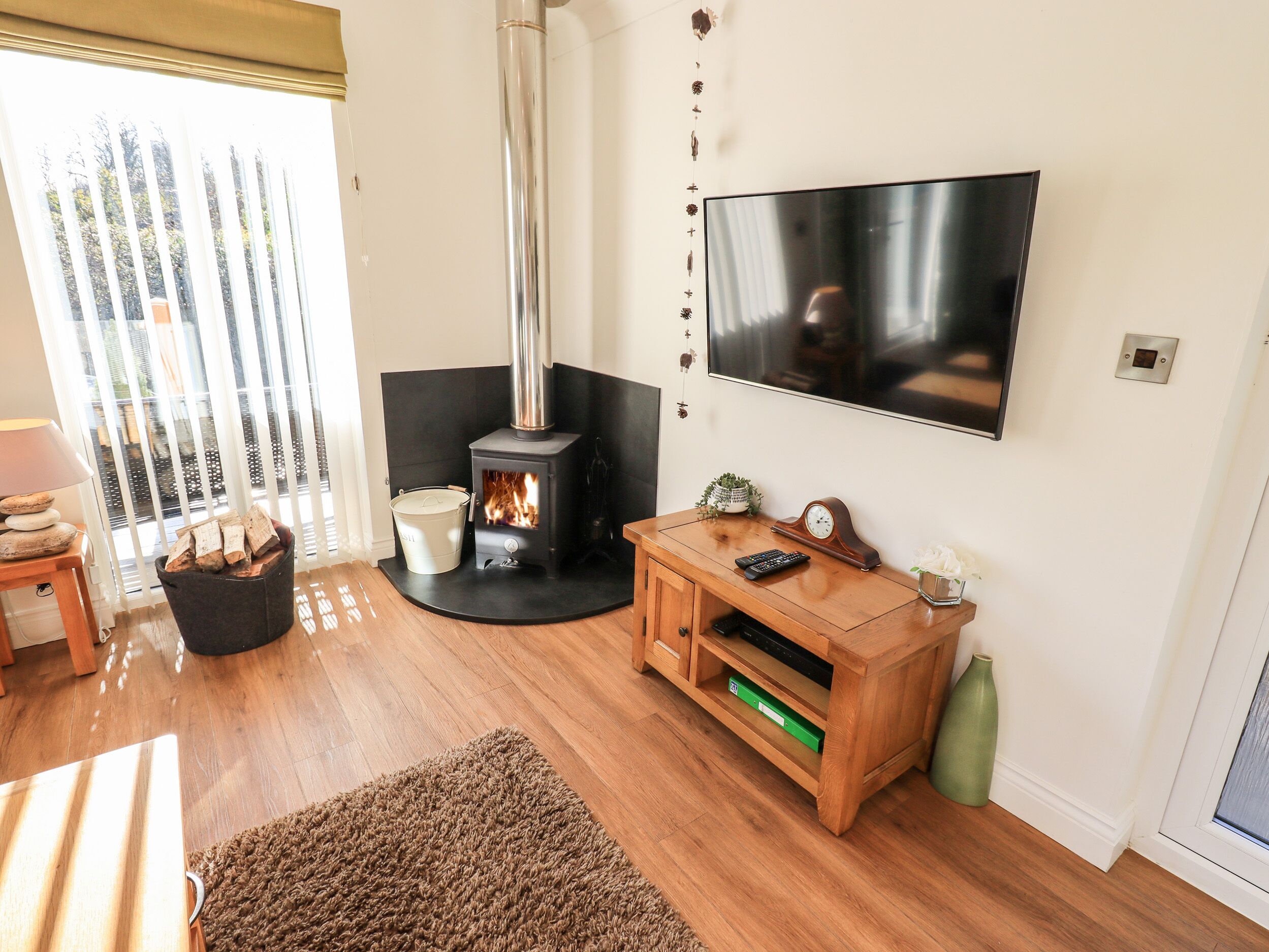 A living room with a wood-burning stove, a TV mounted on the wall, a wooden cabinet, and a large window with blinds.