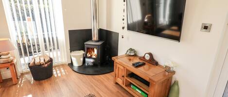 A living room with a wood-burning stove, a TV mounted on the wall, a wooden cabinet, and a large window with blinds.