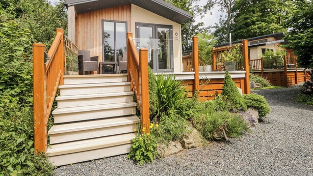 Una cabaña moderna con fachada de madera, puerta de cristal de entrada y una escalera que conduce a una terraza con mobiliario exterior.