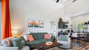 Een moderne woonkamer met een groene bank, een grijze fauteuil en een houten salontafel. Op de achtergrond is een keuken met donkere kasten en een bar te zien.