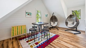 Een knus zolderkamertje met een schuine kap, een ronde tafel met stoelen, twee hangende eierstoelen, een kleurrijk vloerkleed en een prikbord met groene cirkels.