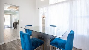 A modern dining area with a glass table and blue chairs, a vase with flowers, and a view through sheer curtains. - Calle Azul in Las Vegas (North Las Vegas)