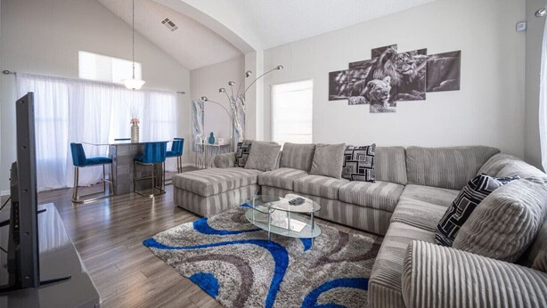 A modern living room with a sectional sofa, a glass coffee table, and a dining area with blue chairs. - Calle Azul in Las Vegas (North Las Vegas)
