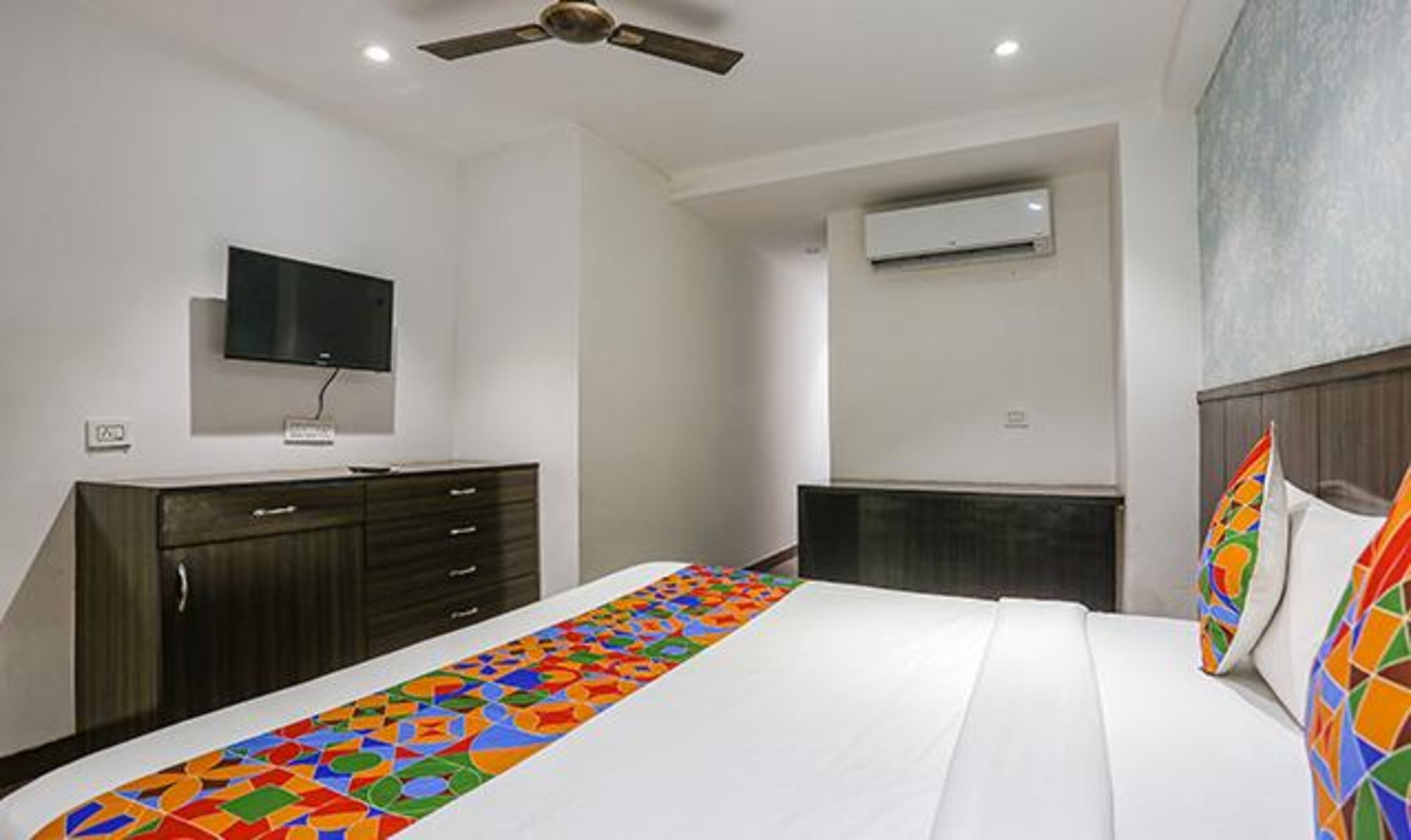 A bedroom with a bed, a TV mounted on the wall, a ceiling fan, and an air conditioning unit.