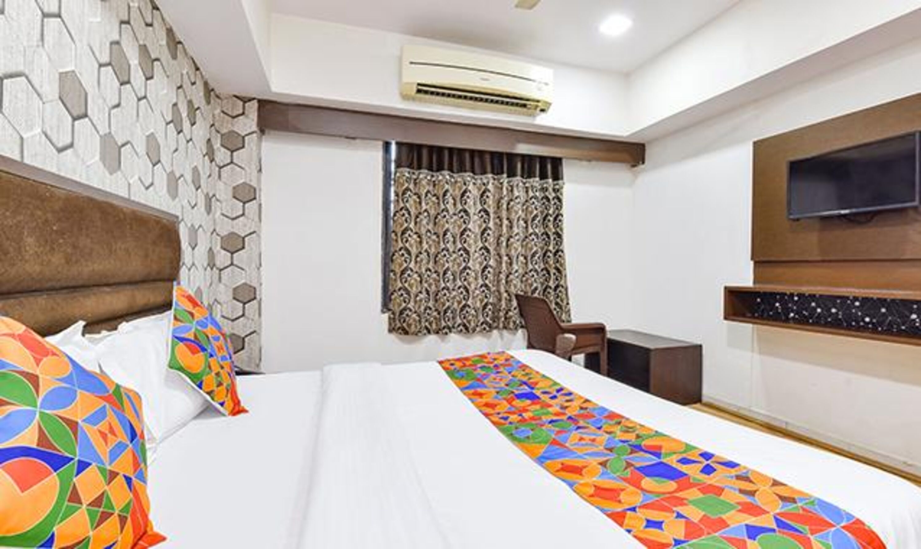A hotel room with a bed, a desk, an air conditioner, a TV, and a patterned curtain.