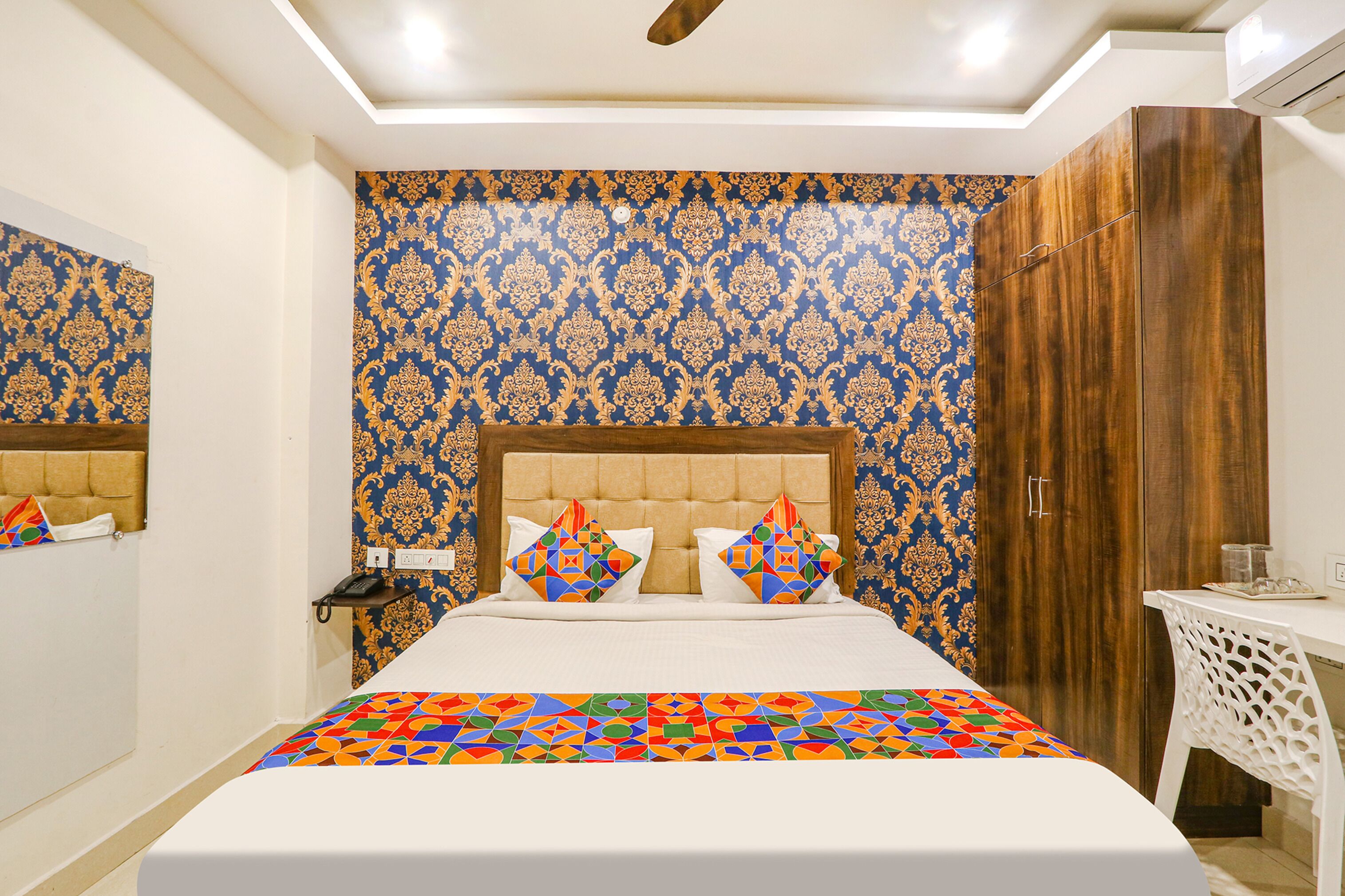 A bedroom with a patterned wallpaper, a tufted headboard, a bed with colorful bedding, a bedside table, and a wardrobe.