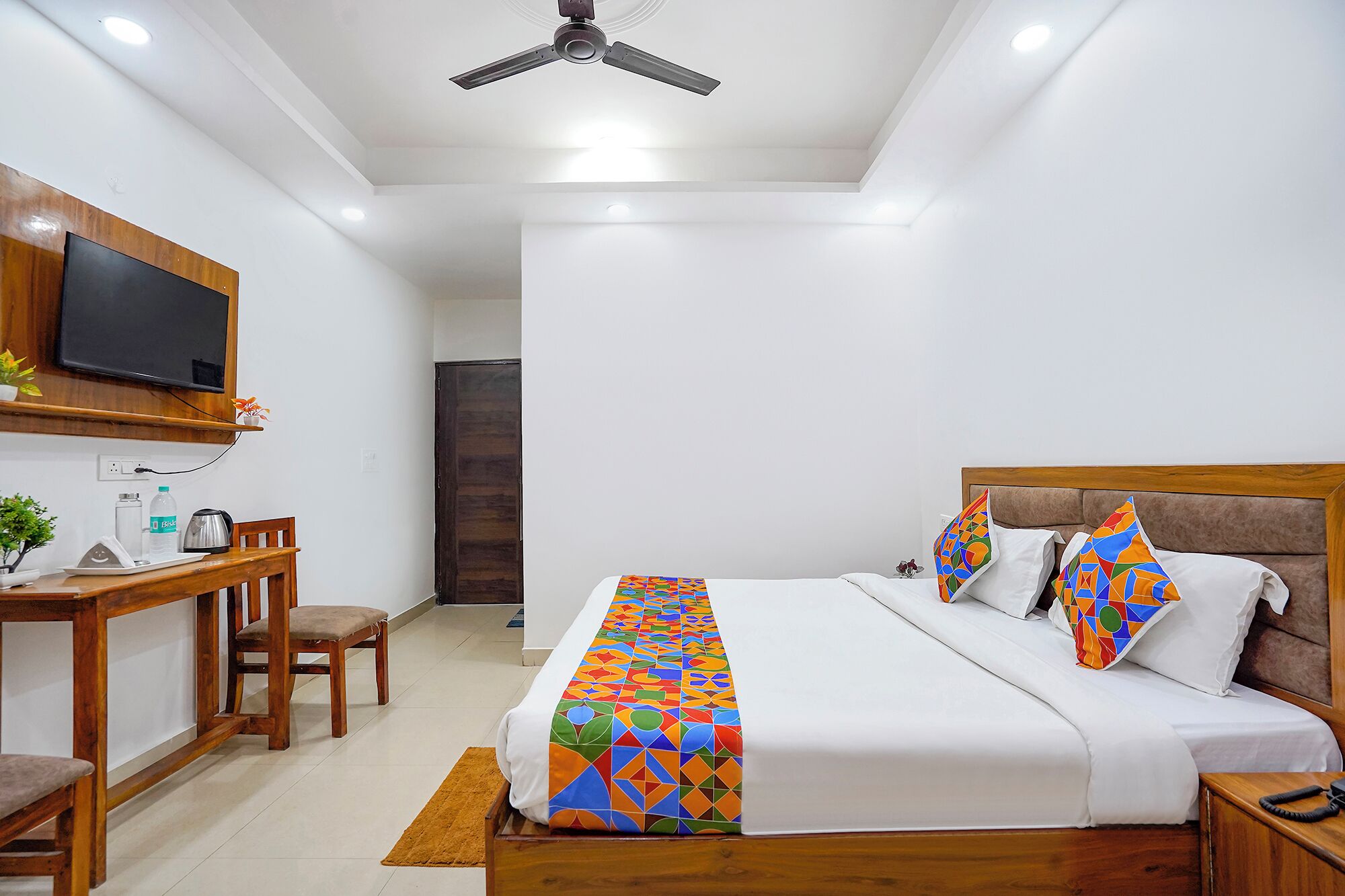 A modern hotel room with a wooden bed, a desk with a chair, a television mounted on the wall, and a ceiling fan.