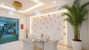 A modern interior with a glass partition, white wicker chairs, a glass table, a large potted plant, and a wall with a floral design. - FabHotel Frolic Luxury Stay (Noida)