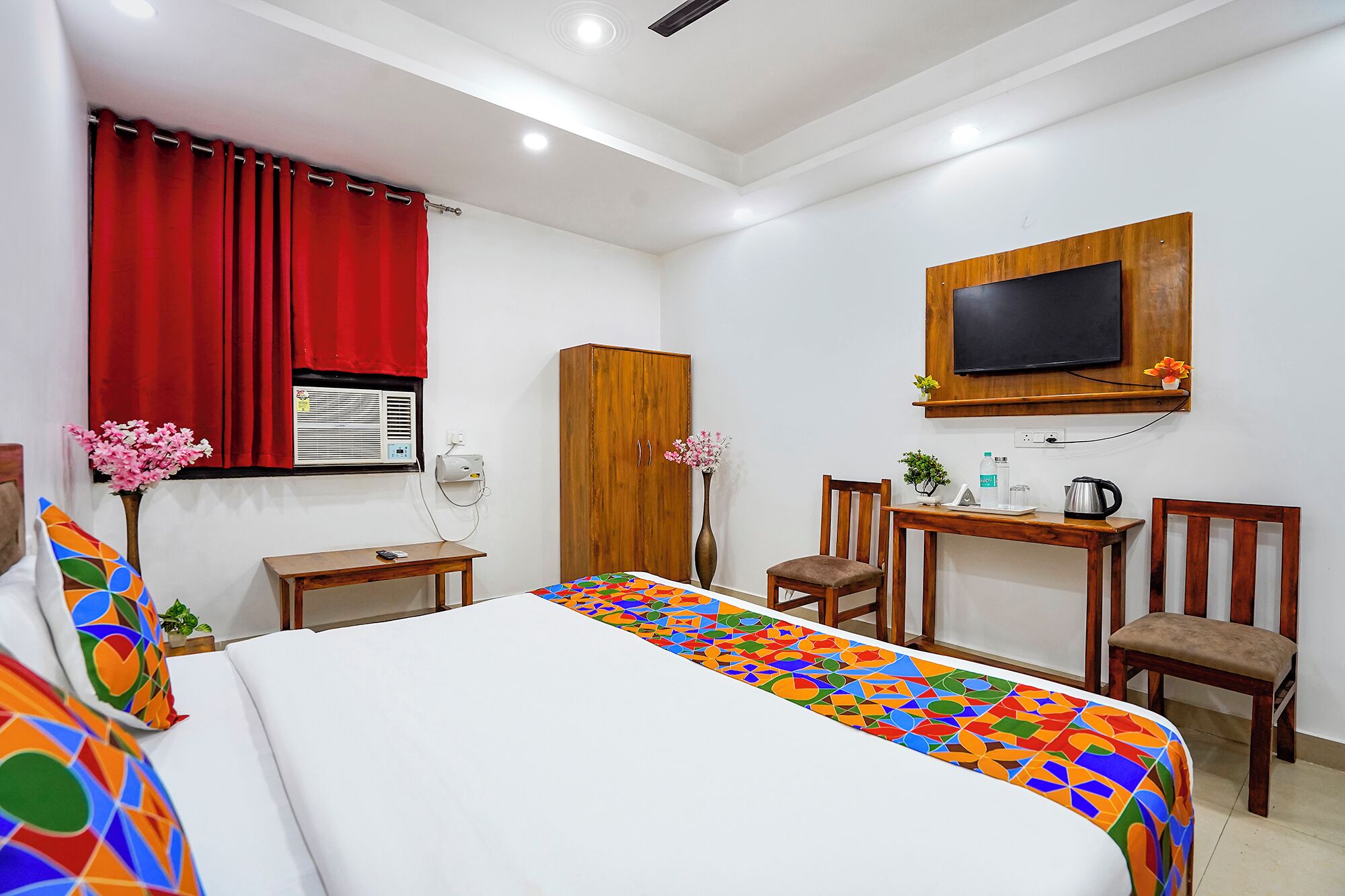 A hotel room with a bed, a desk, a chair, a TV, and a window with red curtains.