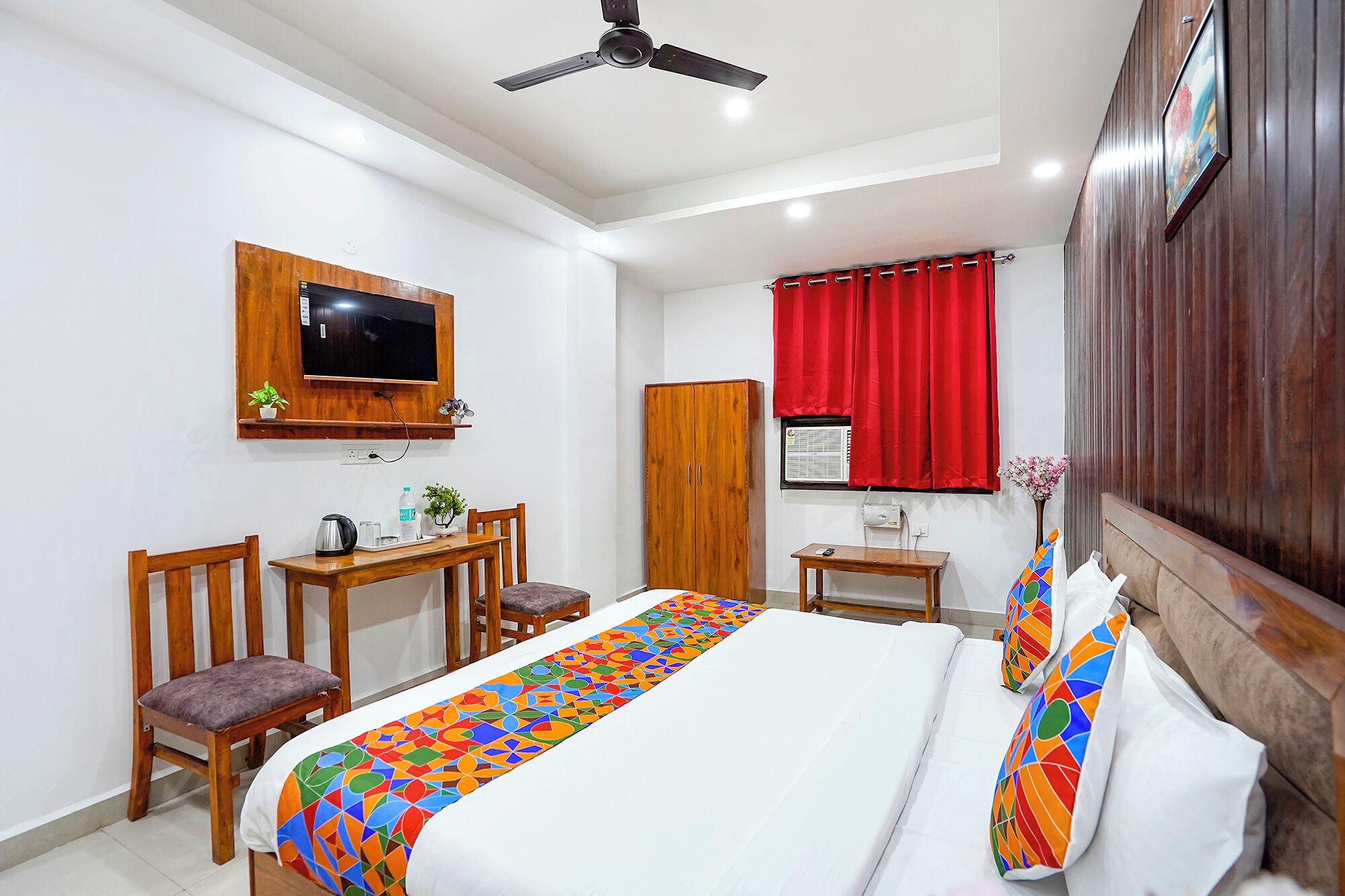 A hotel room with a bed, a desk, two chairs, a television, a wardrobe, and a window with red curtains.