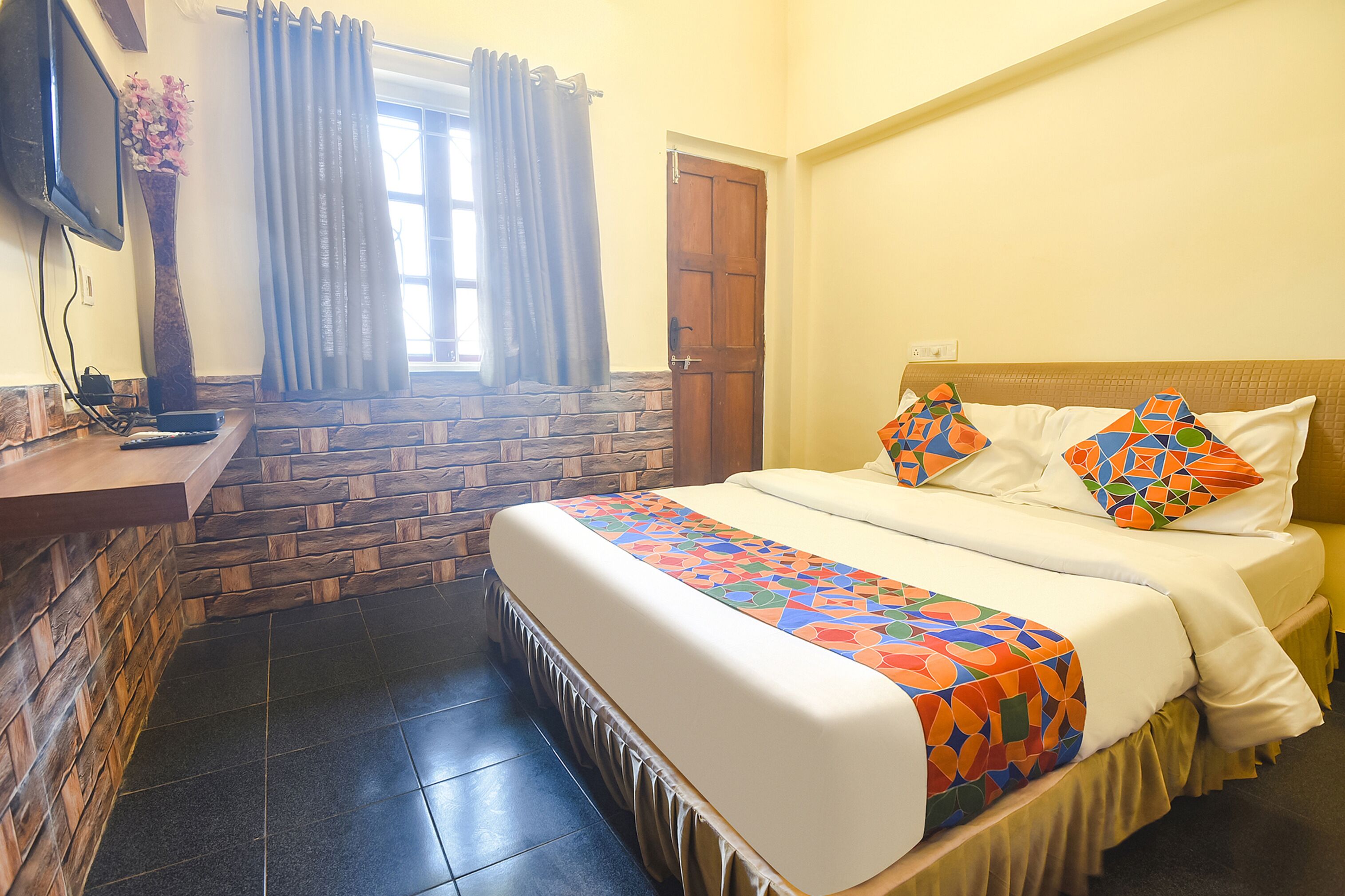 A hotel room with a large bed, a TV, a stone wall, a window with curtains, and a wooden door.