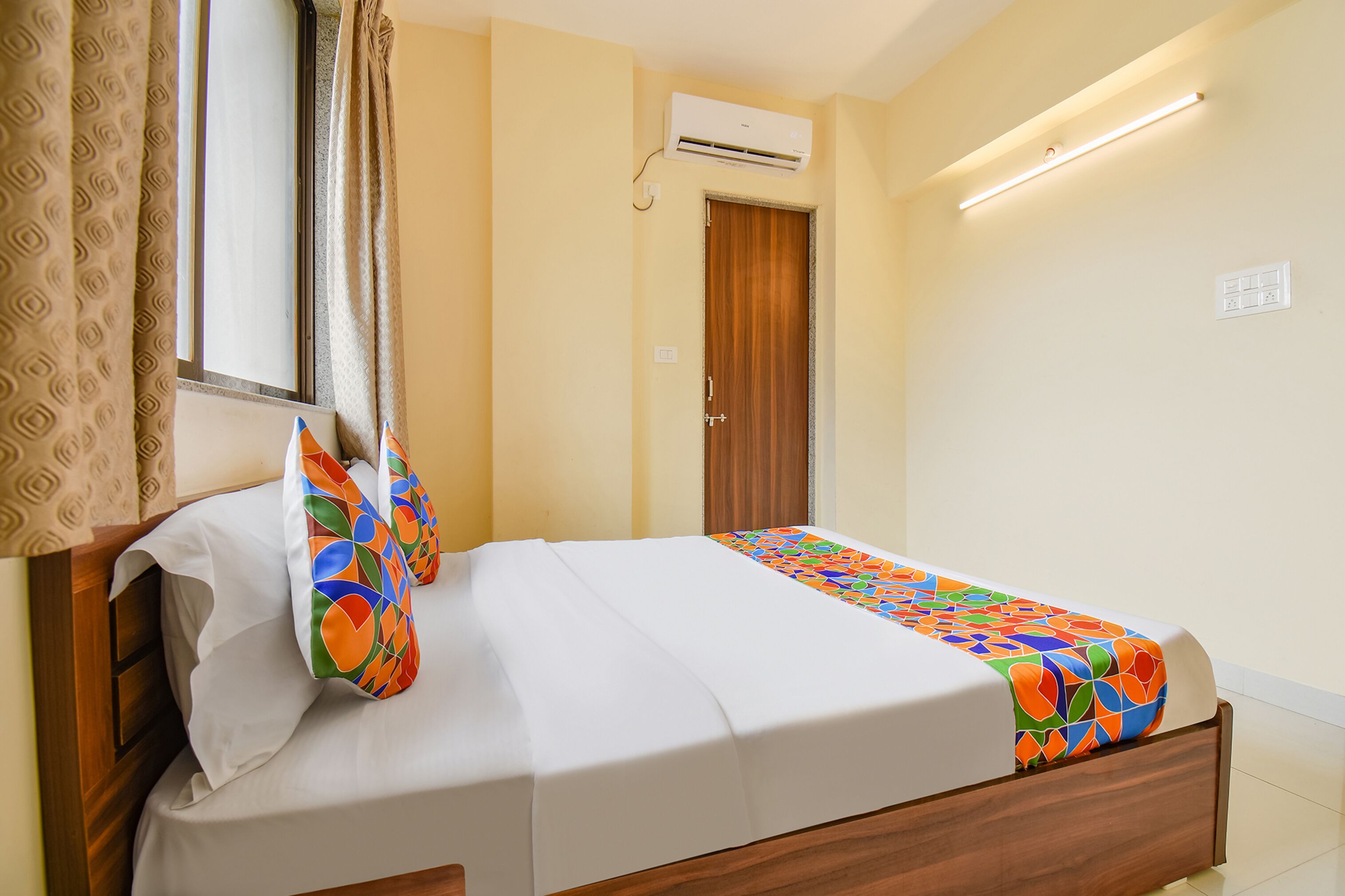 Een net opgemaakt bed met een kleurrijk sprei, een raam met gordijnen, en een airconditioner aan de muur.