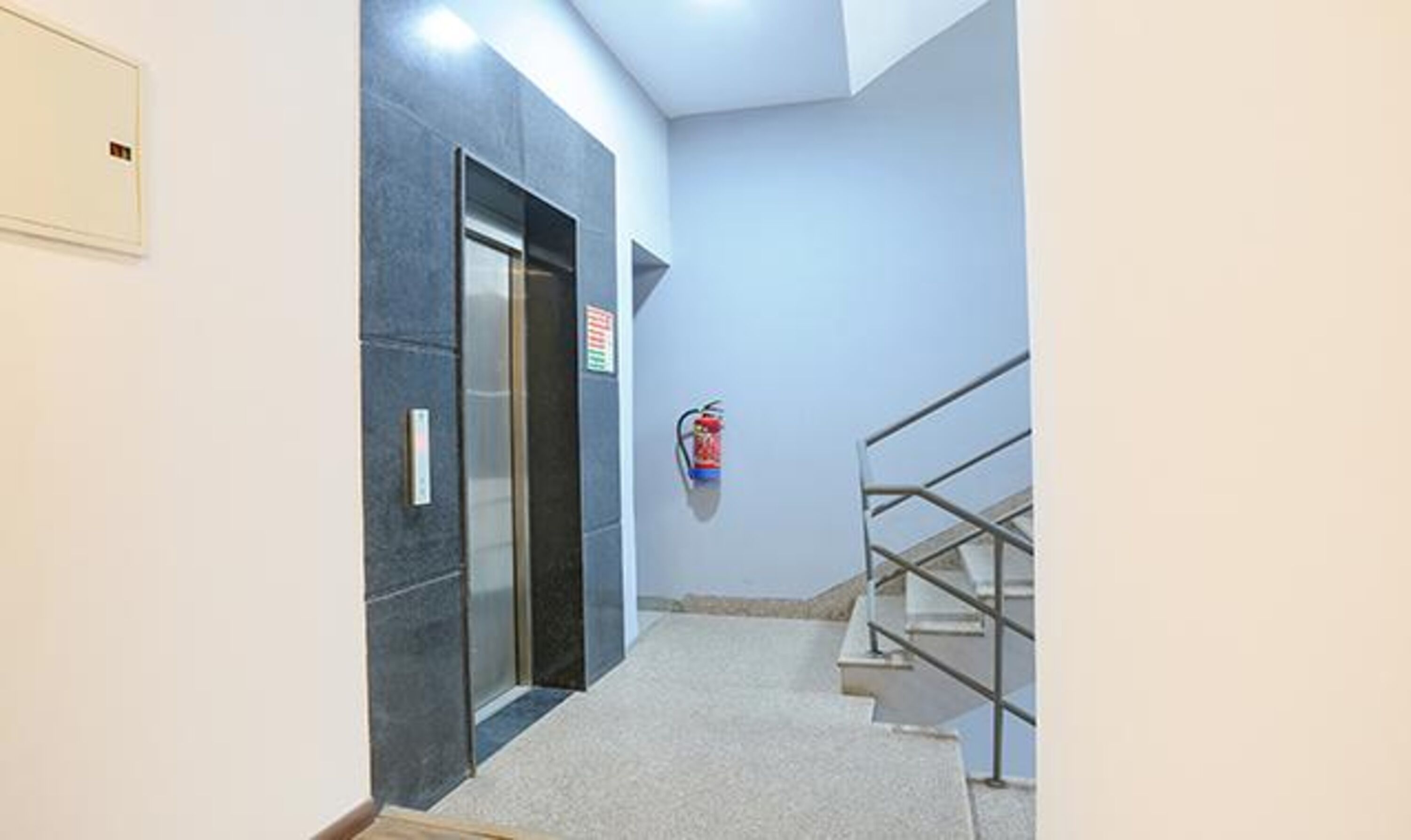 A modern elevator with a digital display, a fire extinguisher mounted on the wall, and a staircase with metal railings.