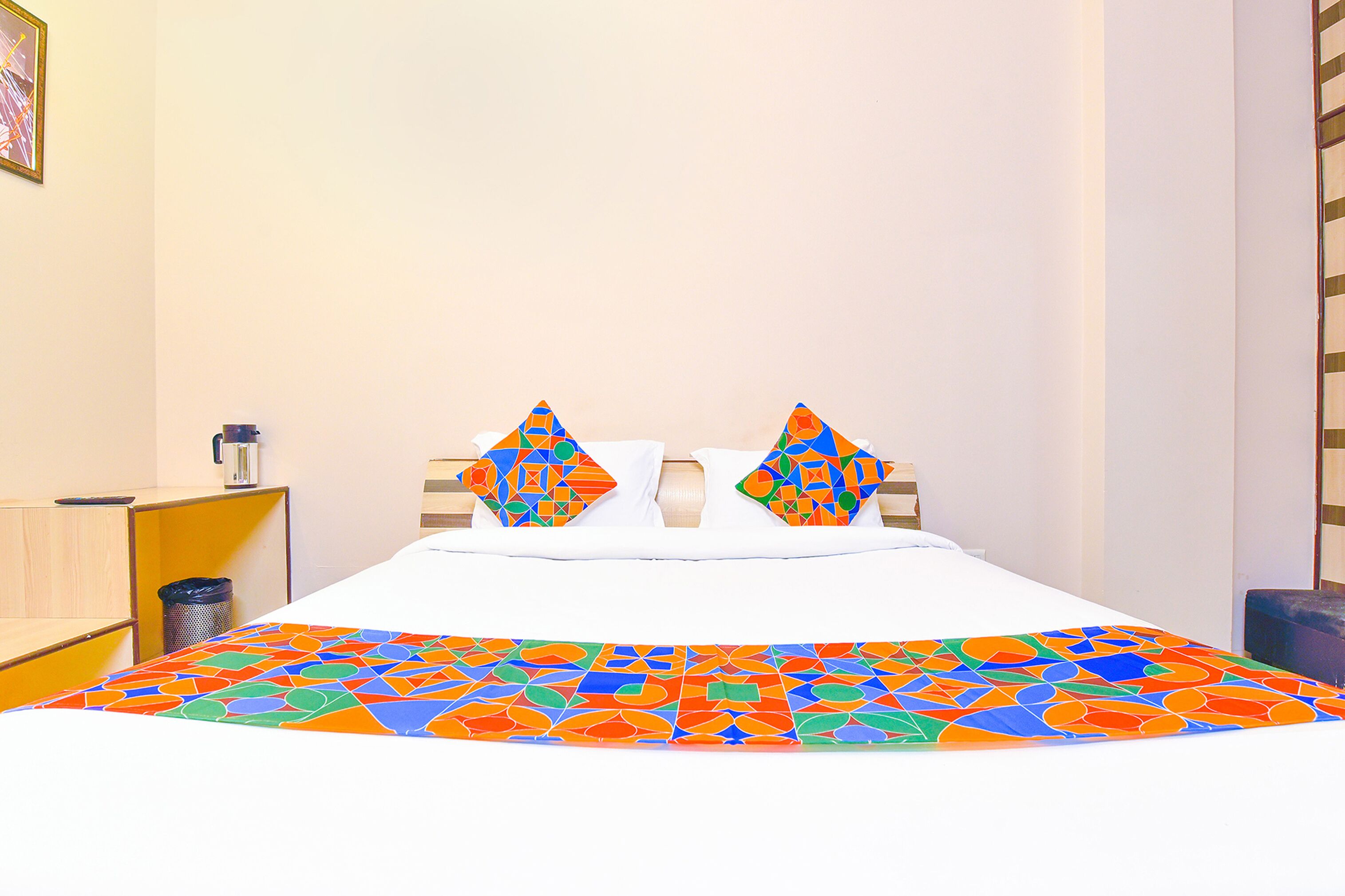 A bed with a colorful geometric-patterned bedspread, two pillows with a similar pattern, and a small trash bin to the left.