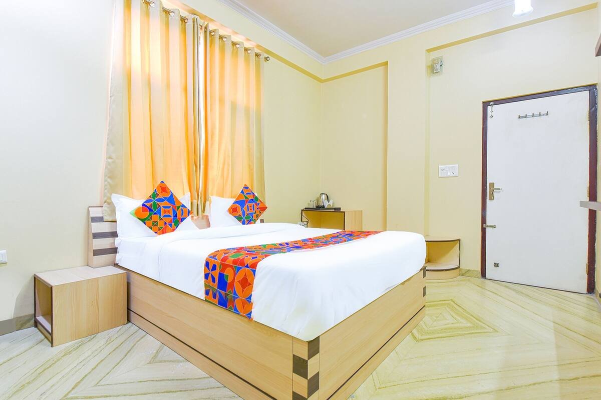 Cama bem arrumada com colcha e travesseiros coloridos, mesa de cabeceira com abajur e porta ao fundo.