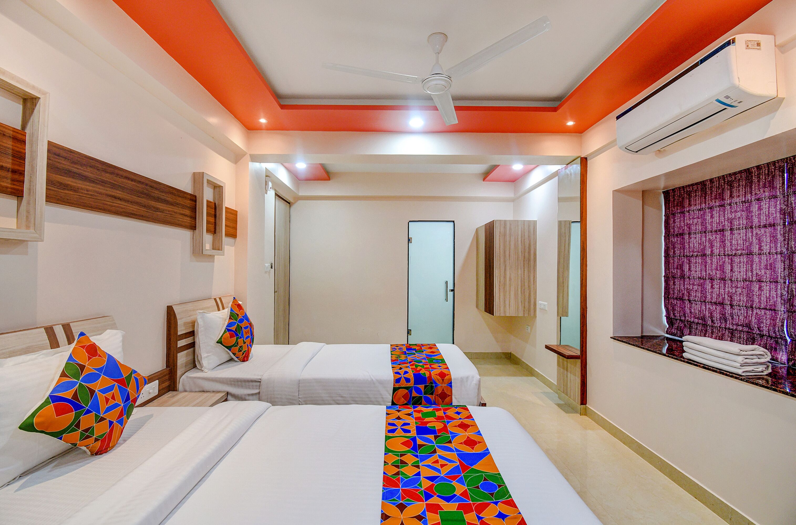A hotel room with two beds, a ceiling fan, an air conditioning unit, a mirror, and a window with curtains.