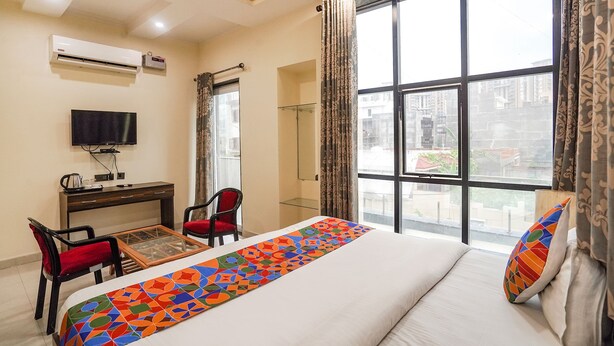 A hotel room with a bed, a desk, a chair, a TV, and a large window with curtains. - Fabhotel 17 Suites (Noida)