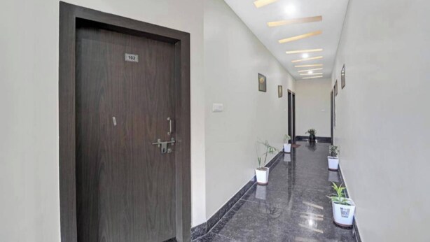 A hallway with a dark wooden door labeled "102", potted plants, and a modern ceiling design. - Hotel Ravi Residency (Bharatpur)