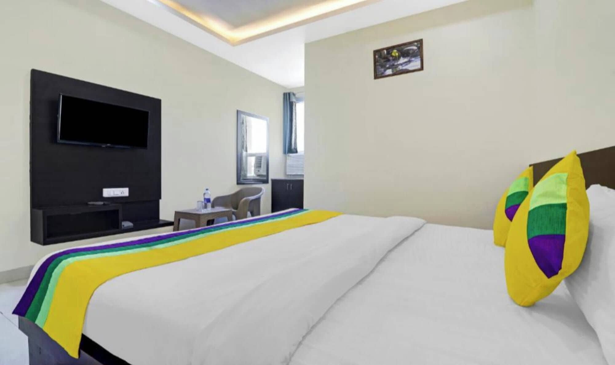 A hotel room with a bed, a TV mounted on the wall, a small table with a chair, and a window with curtains.