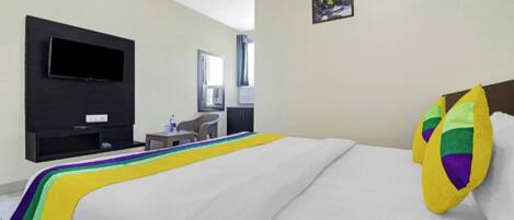A hotel room with a bed, a TV mounted on the wall, a small table with a chair, and a window with curtains.