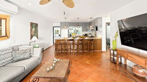 A modern living room with a sofa, coffee table, and a TV. There is a kitchen area with a dining table and chairs.