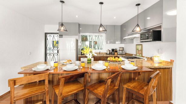 A modern kitchen with a central island, wooden chairs, pendant lighting, and a breakfast setup.