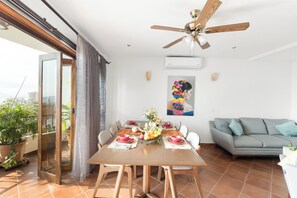 Uma sala de jantar com uma mesa de madeira, cadeiras e um ventilador de teto. Há um sofá cinza ao fundo e uma planta em vaso à esquerda.