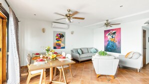 A modern living room with a dining area, two ceiling fans, a grey sofa, a white armchair, and a large painting on the wall.