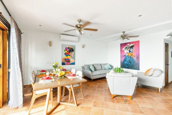 Uma sala de estar moderna com área de jantar, duas ventoinhas de teto, um sofá cinzento, uma poltrona branca e um quadro grande na parede.