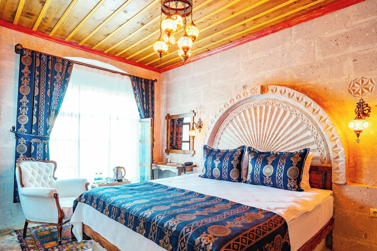 Quarto com cama grande, uma cadeira, escrivaninha e janela com cortinas.