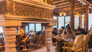Ein Kamin aus Stein mit aufwendigen Schnitzereien, ein Feuer darin und Korbstühle rund um einen Tisch in einem Restaurant.