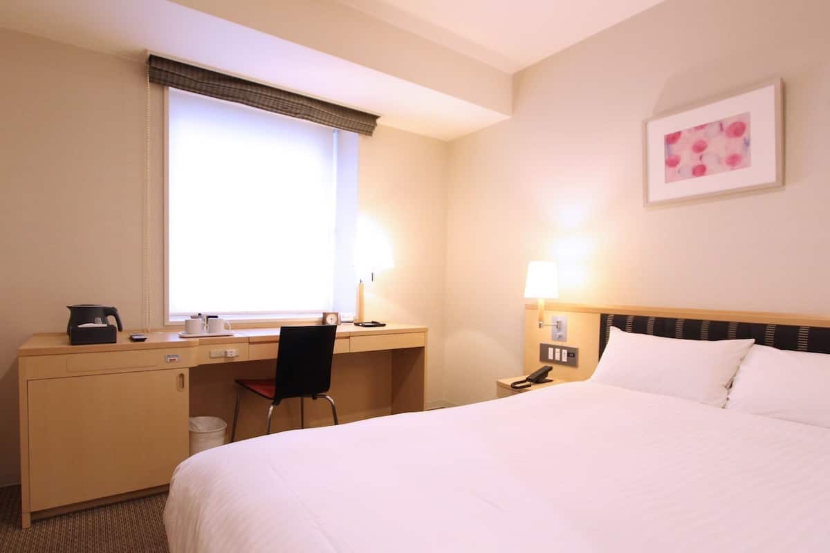 A hotel room with a bed, desk, chair, and a window with blinds.
