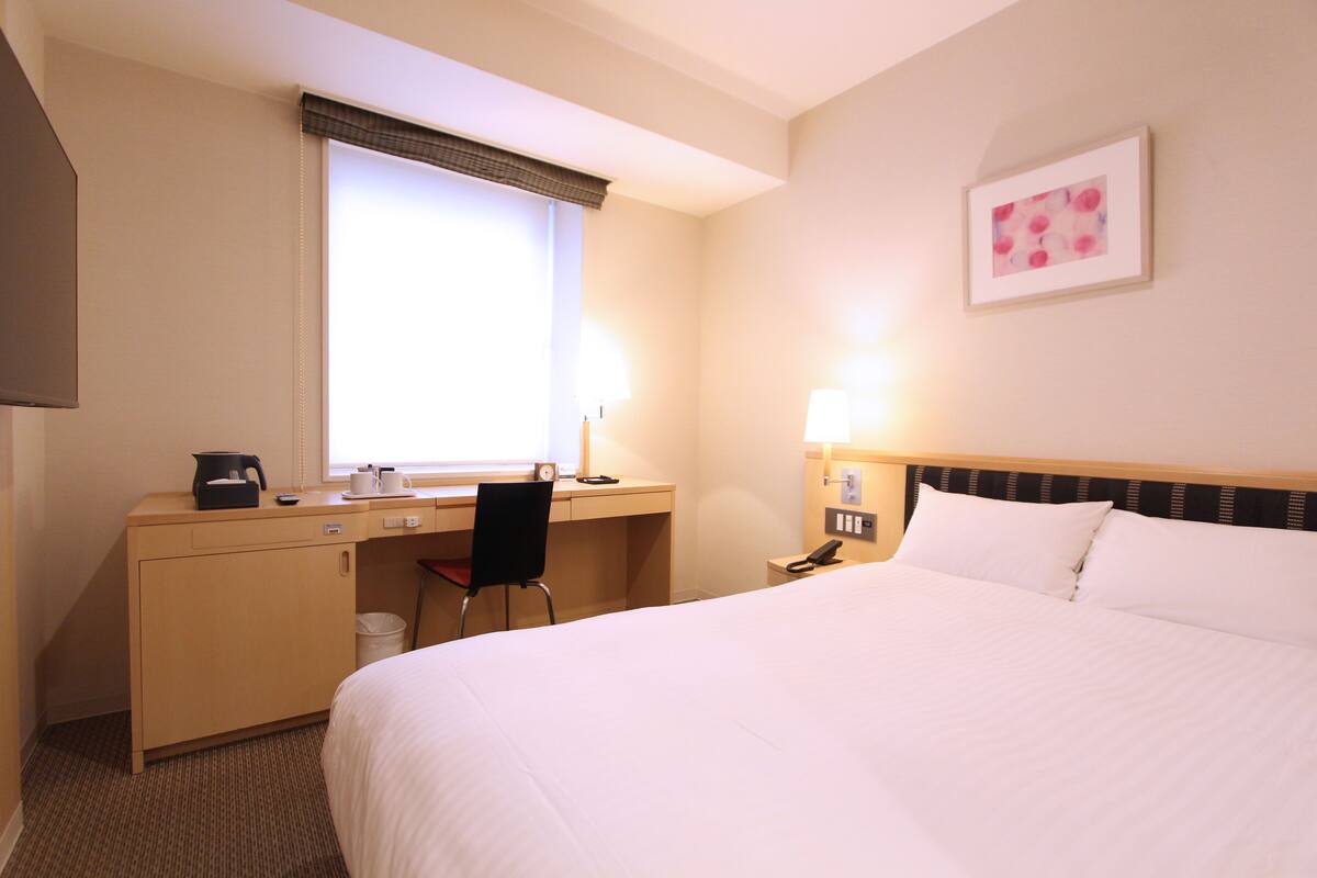 A hotel room with a bed, desk, chair, and a window with blinds.