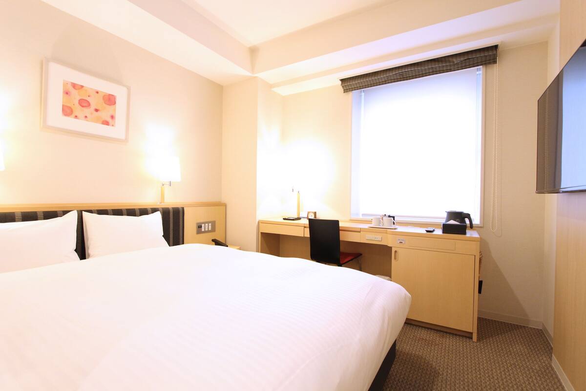 A hotel room with a large bed, a desk, a chair, a television, and a window with blinds.