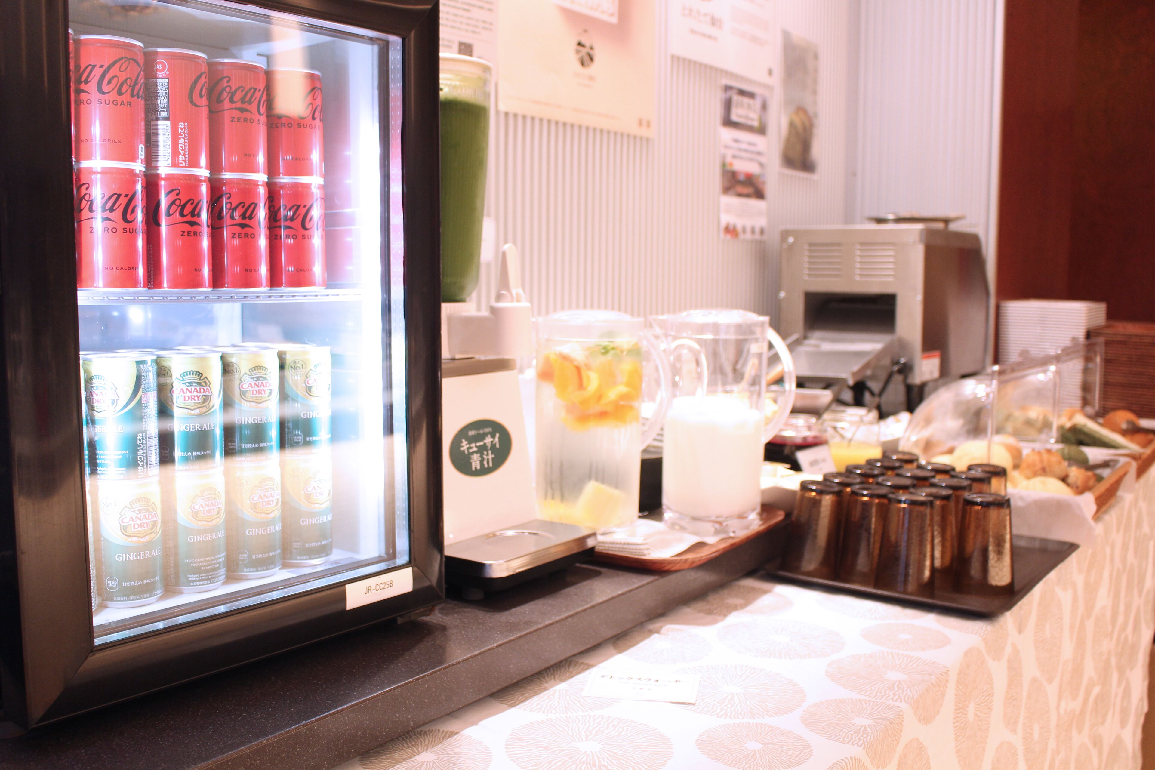 A beverage dispenser with cans of Coca-Cola Zero and cans of ginger ale, a milk dispenser, and a tray with condiments and a pitcher of orange juice.
