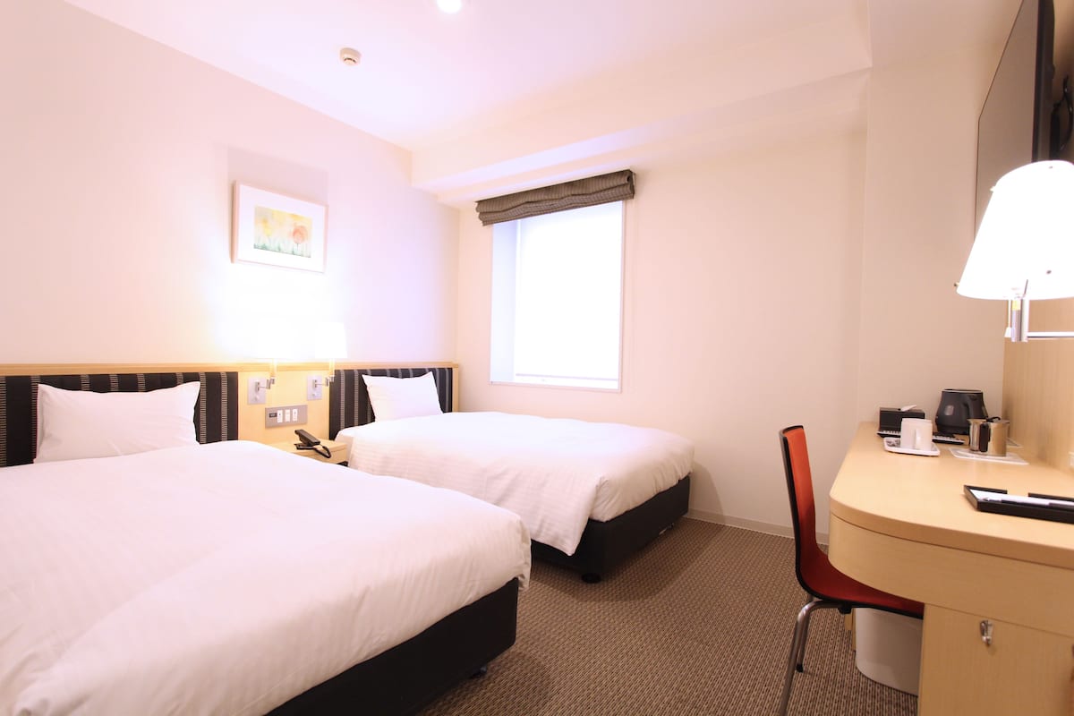 A hotel room with two beds, a desk with a chair, a telephone, and a window with curtains.