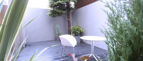 Un patio con piso de madera, dos sillas blancas, una mesita y plantas en macetas.
