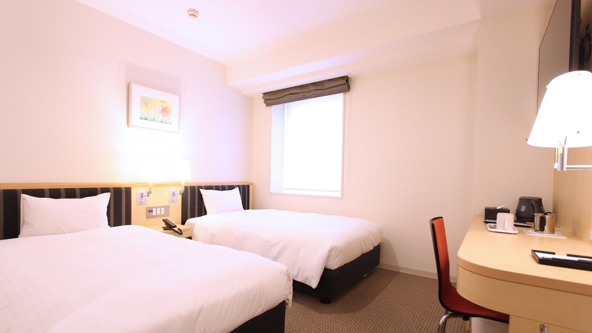 Habitación de hotel con dos camas, un escritorio, una silla, un ventanal con cortinas, una mesita de noche con lámpara y un cuadro en la pared.