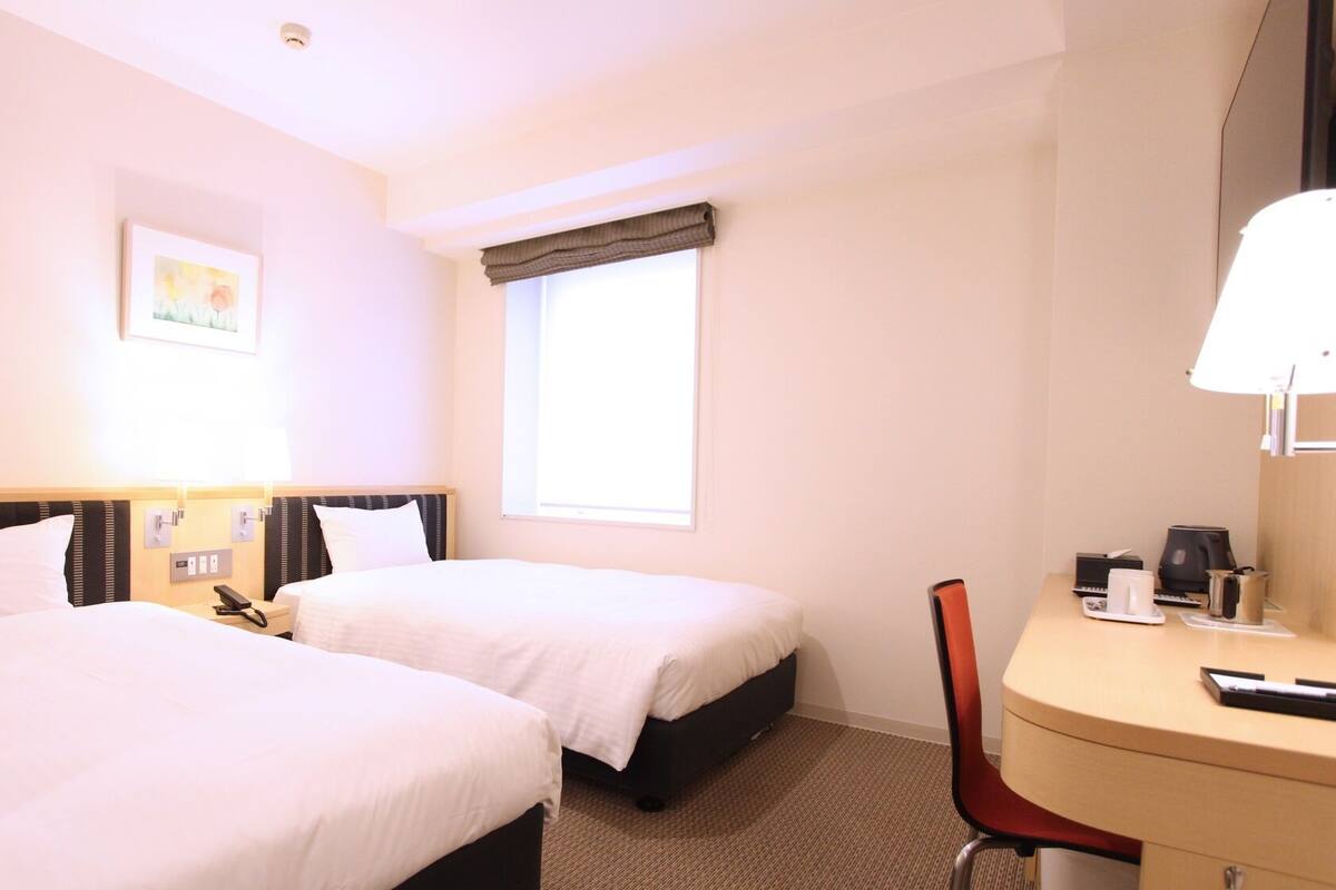 A hotel room with two beds, a desk, a chair, a window with curtains, a bedside table with a lamp, and a painting on the wall.