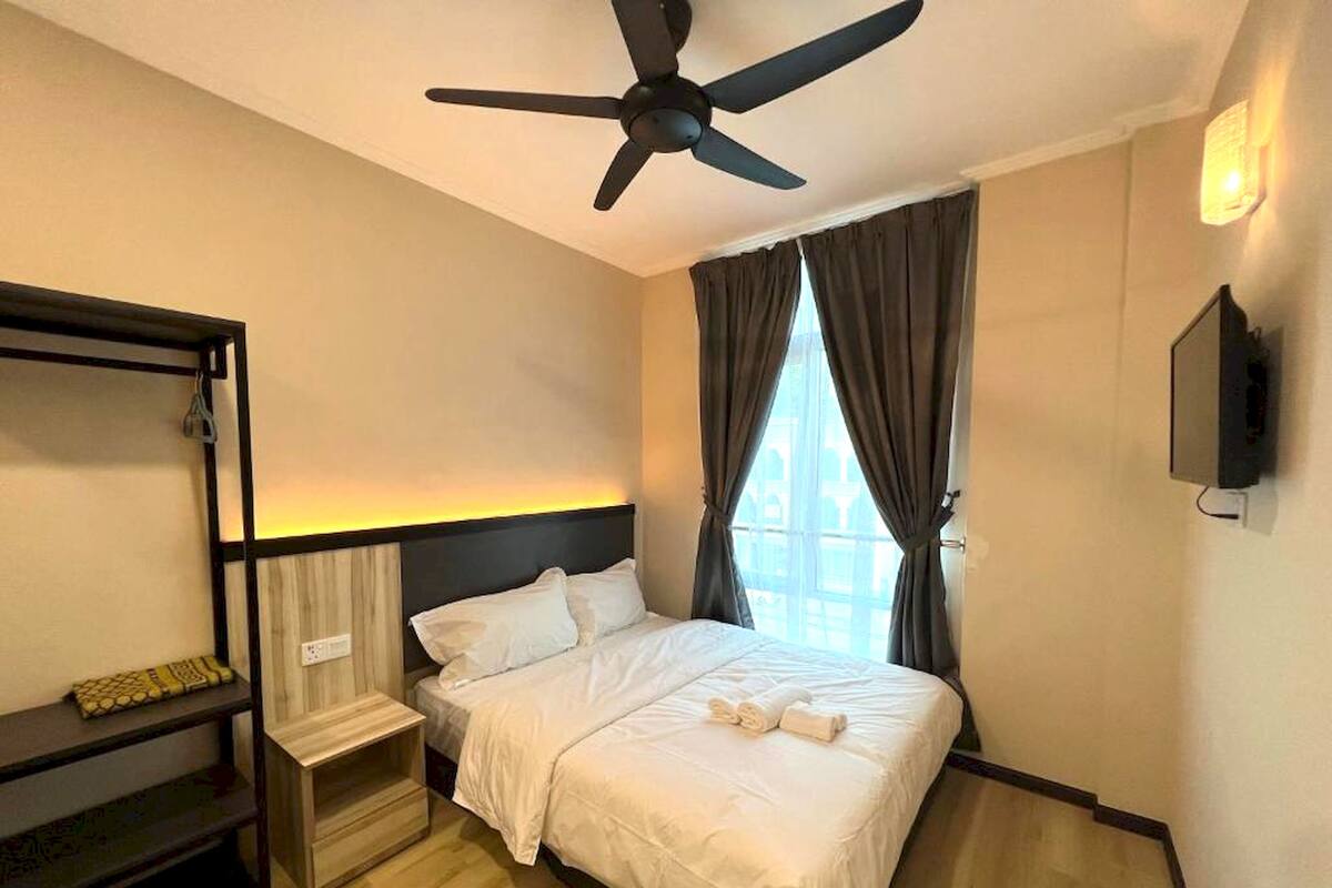 A hotel room with a bed, a ceiling fan, a TV, a window with curtains, and a wooden nightstand.