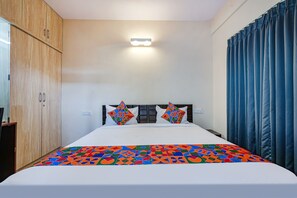 Ein ordentlich bezogenes Bett mit einer farbenfrohen, geometrisch gemusterten Tagesdecke, ein Kopfteil mit zwei dekorativen Kissen, ein Holzschrank und eine wandmontierte Lampe.