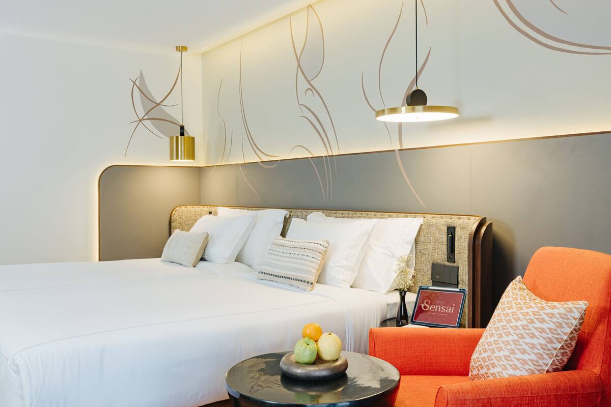 Ein modernes Hotelzimmer mit einem großen Bett, einem orangefarbenen Sessel, einem kleinen runden Tisch mit Früchten und einem Nachttisch mit einem Laptop und einem Schild.