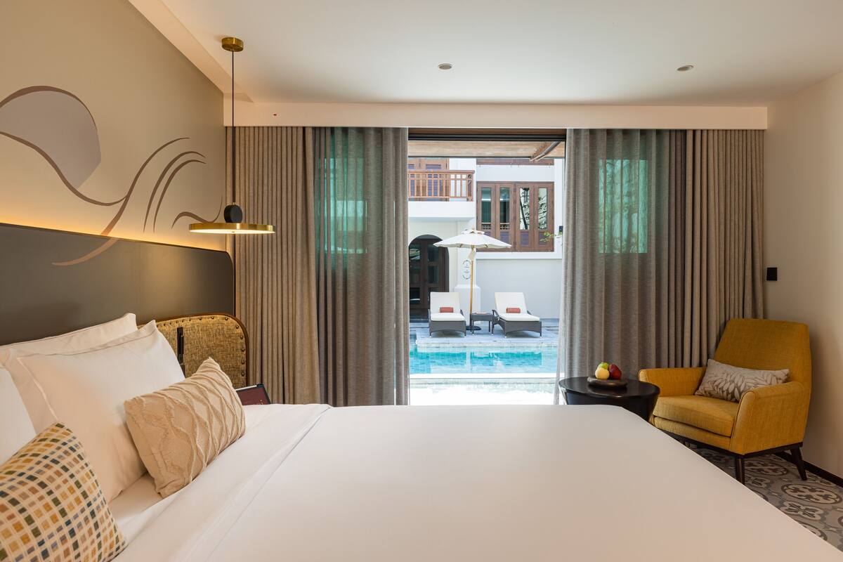 Ein Hotelzimmer mit einem großen Bett, einem kleinen Tisch mit Obstschale, einem gelben Sessel und Blick auf einen Pool und eine Außenbereichs-Sitzgelegenheit.