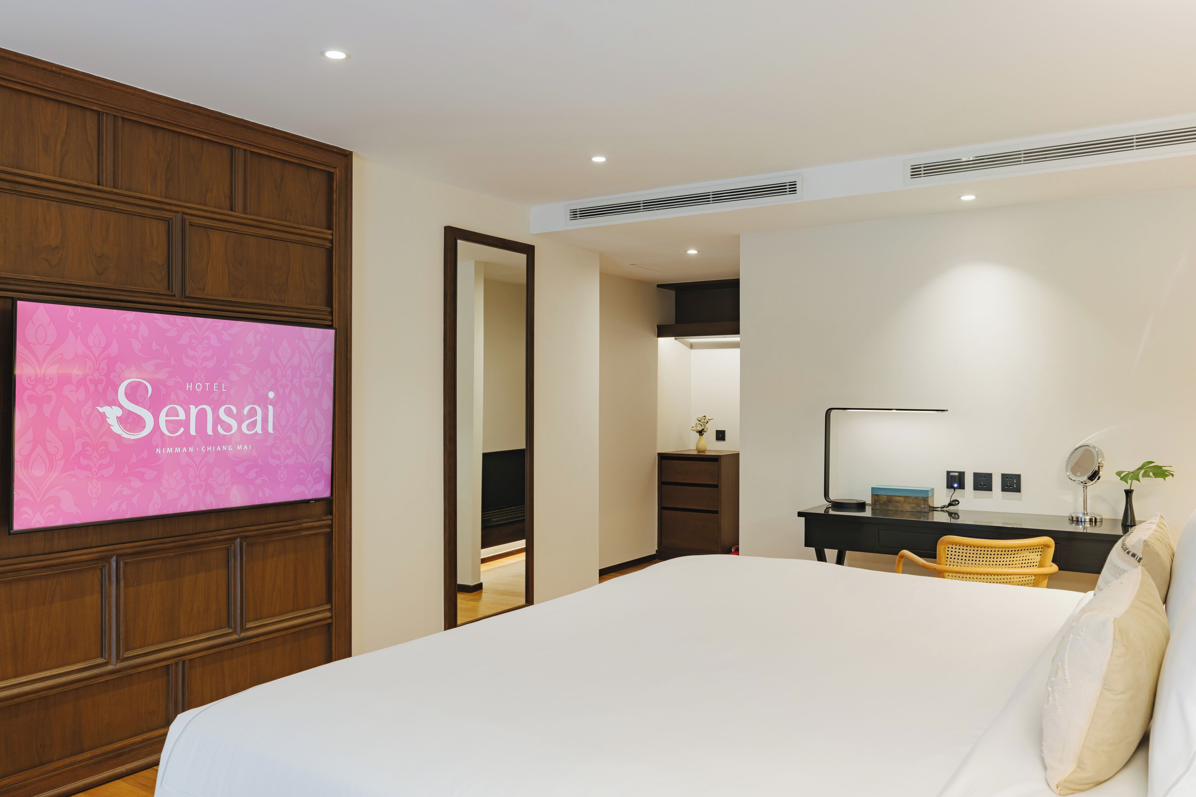 A hotel room with a bed, a desk, a chair, a TV displaying 'Sensai Hotel', and a mirror.