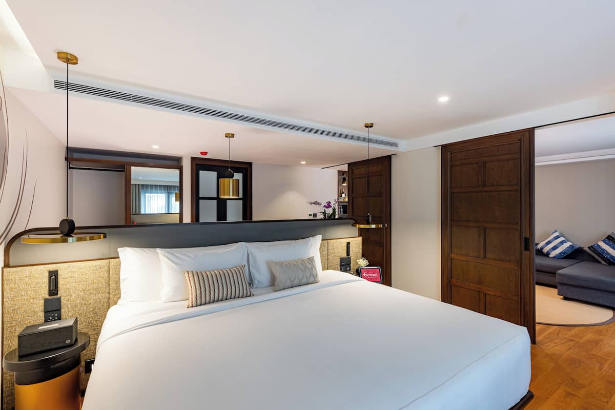 Ein modernes Hotelzimmer mit einem großen Bett, einem Nachttisch, einem Schrank, einem Spiegel und einer Couch im angrenzenden Wohnbereich.
