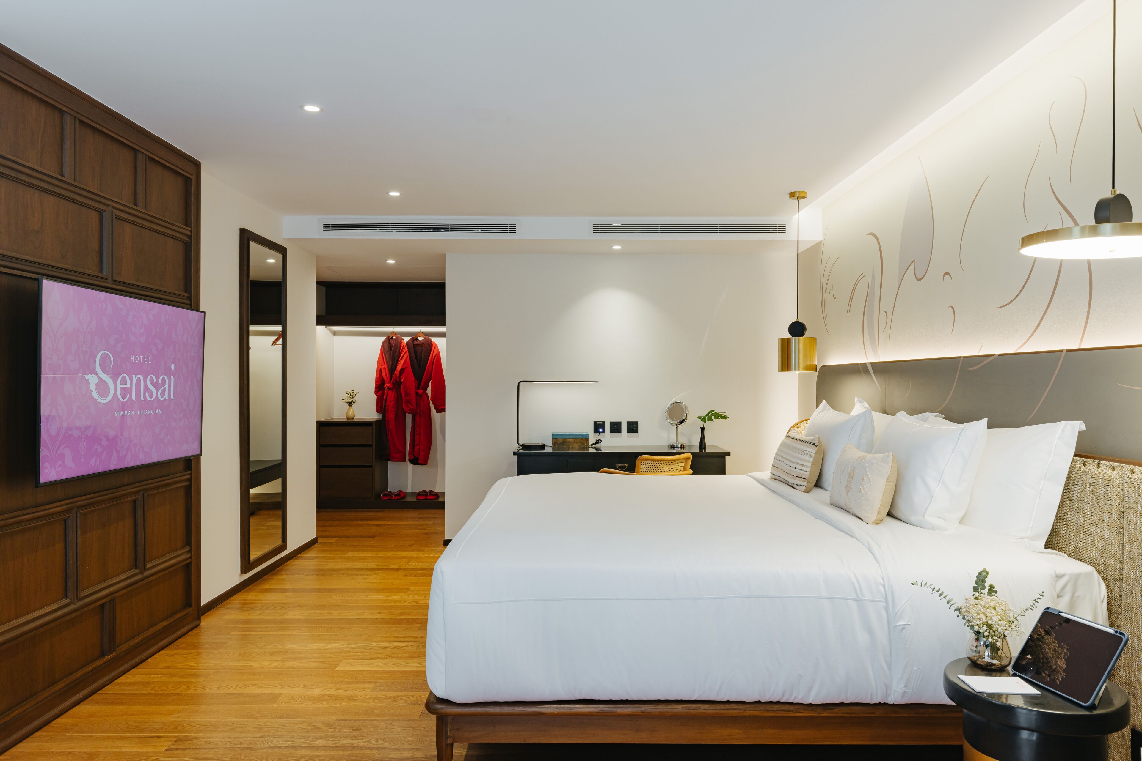Habitación de hotel con una cama grande, una mesita de noche con lámpara, una pequeña mesa con un jarrón y un televisor instalado en la pared.