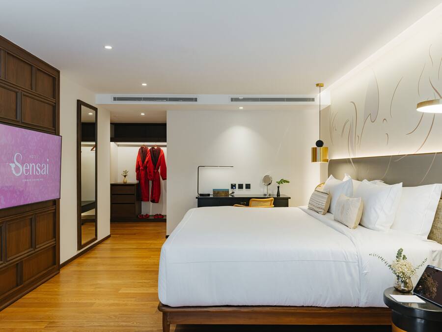 Habitación de hotel con una cama grande, una mesita de noche con lámpara, una pequeña mesa con un jarrón y un televisor instalado en la pared.