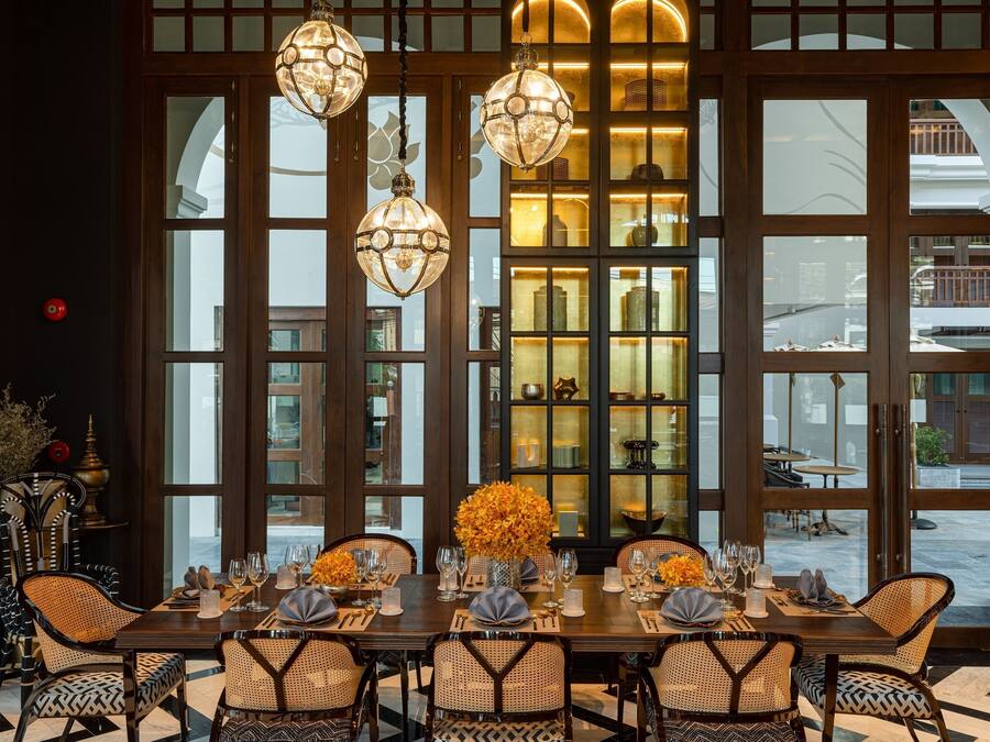 Comedor con una mesa larga dispuesta para seis personas, rodeada de sillas de mimbre, con un ventanal grande y un jarrón decorativo con flores en el centro.